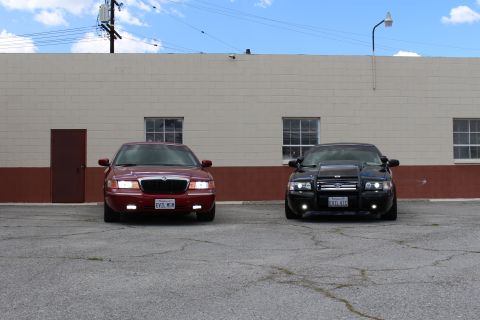 2003 Ford Crown Victoria - maroon Mercury sedan and a black Ford sedan face the camera, parked on cracked pavement before a tan wall.