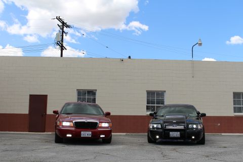 2003 Ford Crown Victoria - maroon Mercury and a black Ford sedan with a grille guard sit front-facing on asphalt below a tall cloudy sky.