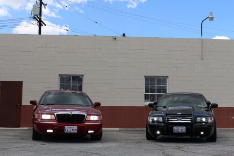 2003 Ford Crown Victoria - maroon Mercury and a black Ford sedan with a grille guard park front-facing before a tan warehouse and power lines.
