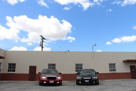 2003 Ford Crown Victoria - Distant front view of a maroon sedan and a black sedan parked together on a cracked lot under a wide blue sky.