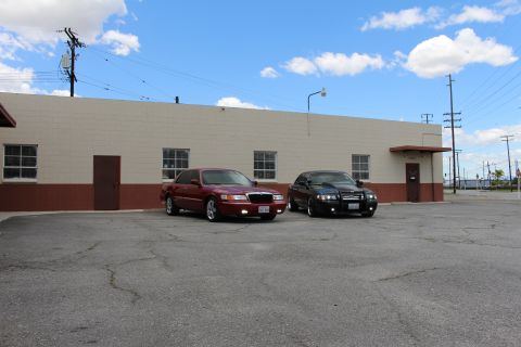 2003 Ford Crown Victoria - maroon Mercury and a black sedan parked at an angle beside a tan warehouse with power poles in the background.