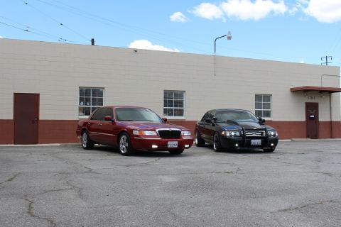 2003 Ford Crown Victoria - Three-quarter view of a maroon Mercury sedan and a black sedan with a grille guard parked before a tan building.