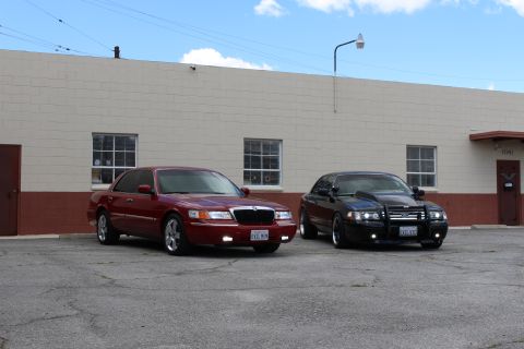 2003 Ford Crown Victoria - maroon Mercury sedan and a black Ford sedan with a push bar sit angled on cracked asphalt by a tan warehouse.