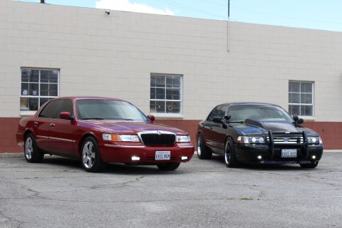 2003 Ford Crown Victoria - maroon Mercury sedan beside a black Ford sedan with a grille guard, both angled on cracked asphalt by a building.