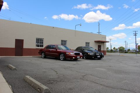 2003 Ford Crown Victoria - Wide shot of a maroon Mercury and a black sedan parked side by side along a tan warehouse with a parking lot beyond.