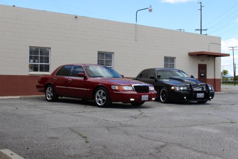 2003 Ford Crown Victoria - maroon Mercury sedan and a black Ford sedan park angled on a broad cracked lot beside a tan cinder-block building.