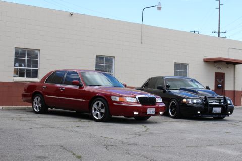 2003 Ford Crown Victoria - maroon Mercury sedan on polished wheels beside a black Ford sedan with a grille guard, parked by a tan warehouse.