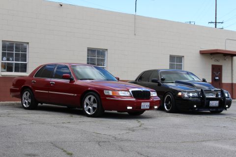 2003 Ford Crown Victoria - maroon Mercury sedan and a black Ford sedan with a push bar parked at an angle on cracked asphalt by a building.