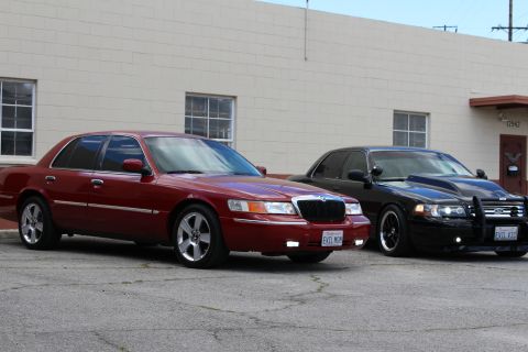2003 Ford Crown Victoria - Front-side view of a maroon Mercury EVIL MOM and a black Ford EVIL VIC with grille guard parked before a tan wall.