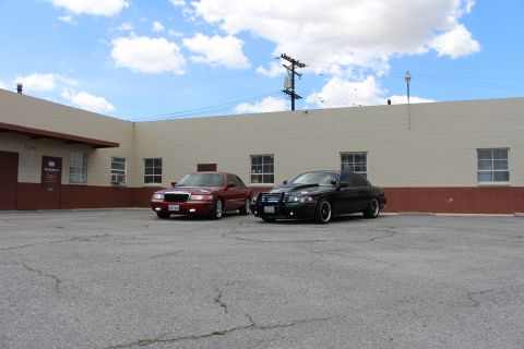 2003 Ford Crown Victoria - Maroon Mercury and black sedan parked together against a tan warehouse under a blue sky with scattered clouds.