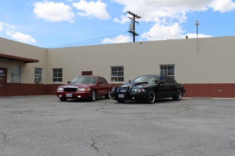 2003 Ford Crown Victoria - maroon Mercury and a black Ford sedan parked angled in a lot beside a tan warehouse with a power pole behind.
