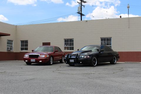 2003 Ford Crown Victoria - maroon Mercury sedan and a black Ford sedan with a grille guard parked at an angle on a cracked lot by a building.