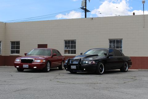 2003 Ford Crown Victoria - Front-side view of a maroon Mercury sedan and a black Ford sedan with a push bar parked before a tan warehouse.