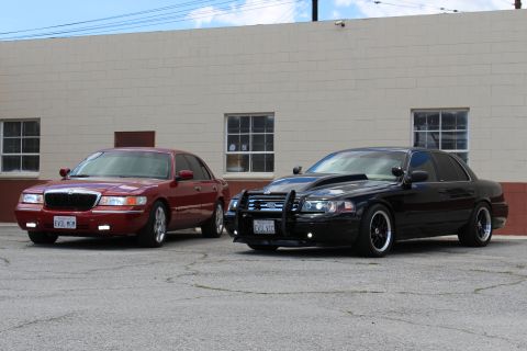 2003 Ford Crown Victoria - maroon Mercury EVIL MOM sedan beside a lowered black Ford with a grille guard parked angled before a tan wall.