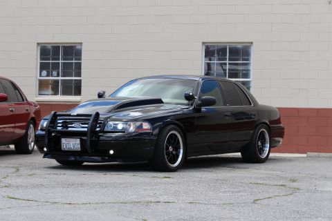 2003 Ford Crown Victoria - lowered black Ford sedan fitted with a black push bumper and black multi-spoke wheels sits in a lot, wearing a California EVIL VIC plate.