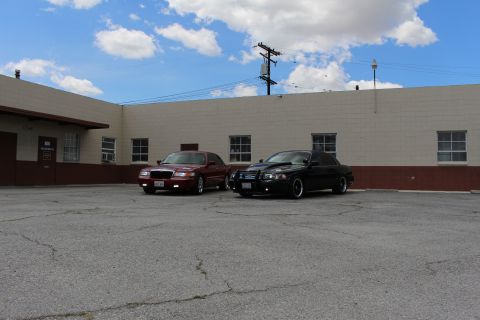 2003 Ford Crown Victoria - dark red sedan and a black sedan sit side by side in a cracked parking lot under a blue sky in front of a tan warehouse.