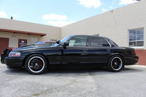 2003 Ford Crown Victoria - lowered black Ford sedan shown in side profile, fitted with black wheels and a front push bumper, parked beside a building.