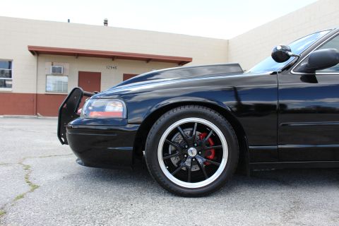2003 Ford Crown Victoria - Close-up of a black sedan's front corner showing a black multi-spoke wheel, red brake caliper, headlight and push bumper.