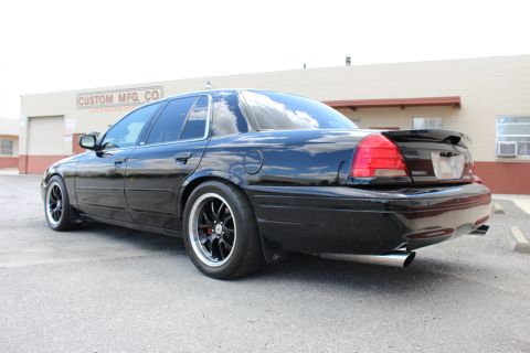 2003 Ford Crown Victoria - Rear three-quarter view of a lowered black sedan with black wheels parked in front of a building marked Custom Mfg Co.