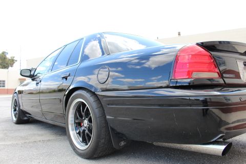 2003 Ford Crown Victoria - Low rear-quarter shot of a black sedan showing its red taillight, black wheel and chrome dual exhaust tip against a cloudy sky.