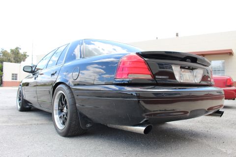 2003 Ford Crown Victoria - Rear three-quarter view of a black sedan with a trunk spoiler and black wheels parked in a lot beside a building.