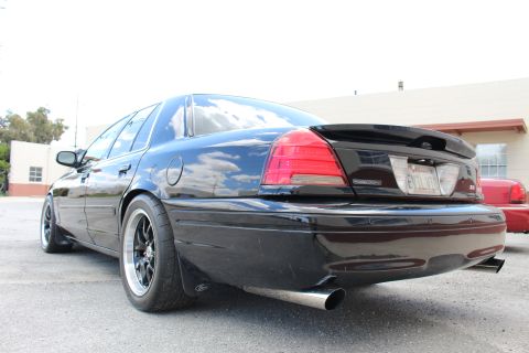 2003 Ford Crown Victoria - Rear three-quarter view of a black sedan with a trunk spoiler, EVIL VIC-style plate and black wheels in a parking lot.