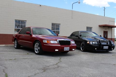 2003 Ford Crown Victoria - dark red Mercury sedan and a black Ford sedan with push bumper sit angled side by side in front of a tan building.
