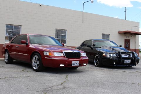 2003 Ford Crown Victoria - dark red Mercury and a black Ford sedan with a push bumper park side by side, both wearing EVIL-themed California plates.