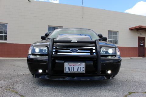 2003 Ford Crown Victoria - Head-on view of a black Ford sedan with halo headlights, fog lights, black push bumper and a California EVIL VIC plate.