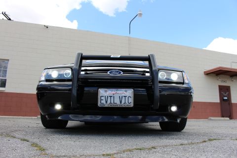 2003 Ford Crown Victoria - Low-angle head-on view of a black Ford sedan with a push bumper, halo and fog lights, and a California EVIL VIC plate.