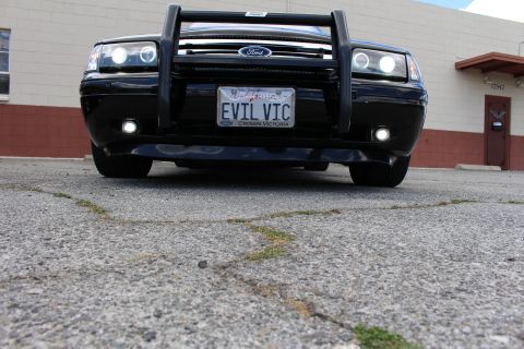 2003 Ford Crown Victoria - Low-angle head-on view of a black Ford sedan with a push bumper, halo lights and a California EVIL VIC plate on cracked pavement.