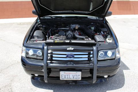 2003 Ford Crown Victoria - Engine bay view of a black Ford sedan with a front brush guard, halo headlights and a California 'EVIL VIC' license plate.