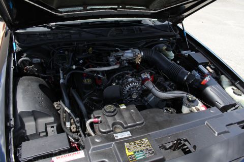 2003 Ford Crown Victoria - Close engine bay view of a dark sedan showing the alternator, ribbed air intake tube and battery with a warning label.