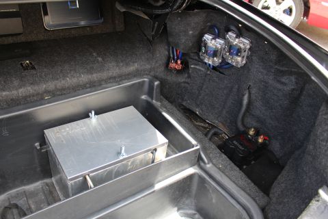 2003 Ford Crown Victoria - Corner of a car trunk showing an aluminum battery box, clear-cased fuse holders and a ground terminal mounted on carpet.