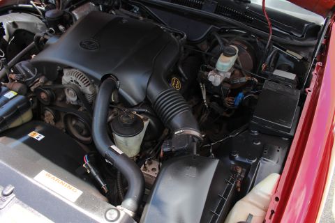 2003 Ford Crown Victoria - Close-up of a red car's engine bay with a black engine cover, alternator and ribbed air intake tube above a warning label.