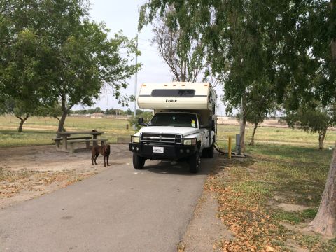 1997 Dodge 3500 - A white pickup truck with a camper shell is parked at a campsite, with a dog standing to its left.