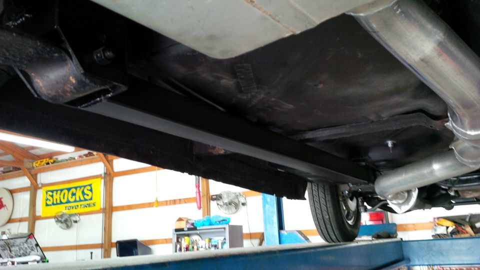 Underside view of a car showing the frame, exhaust pipes, and a rear wheel. A 'SHOCKS TOYO TIRES' sign is visible in the background
