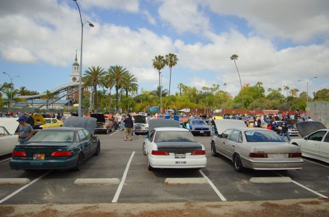 1995 Ford Taurus - A gathering of cars in a parking lot under a partly cloudy sky, with palm trees and a building in the background.