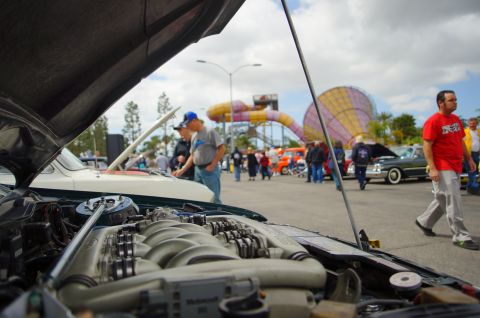 1995 Ford Taurus - A car show features various vehicles and attendees under a cloudy sky with a colorful amusement park structure in the distance.