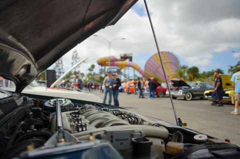 1995 Ford Taurus - A car show features multiple vehicles with hoods up, engines and gathering crowds under a cloudy sky.