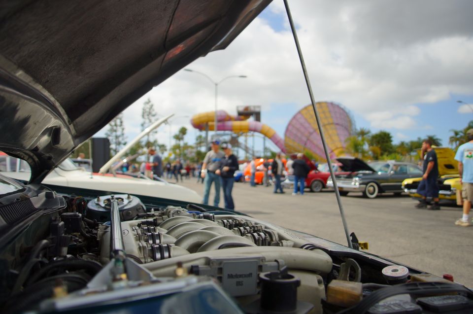 close-up of a car engine with hood open, people and other cars in background at an outdoor event