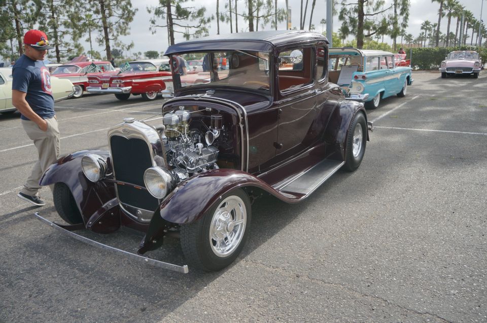 Hot rod coupe with exposed V8 engine, chrome grille, and aftermarket wheels. Other classic cars are parked in the background