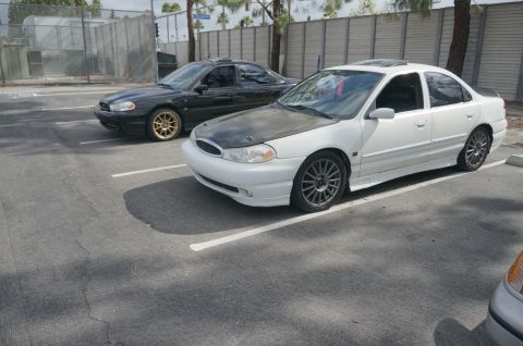 1995 Ford Taurus - Two sedans, a white one with a modified hood and a black one, are parked side-by-side in a lot.