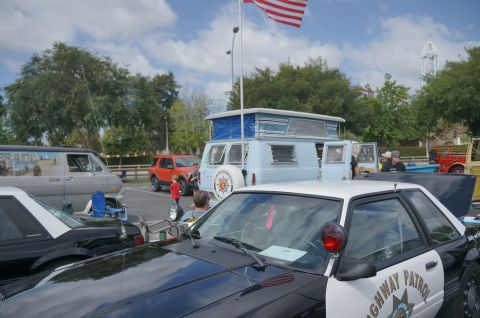 1995 Ford Taurus - A police car is parked at an outdoor event with other vehicles and an American flag flying overhead.