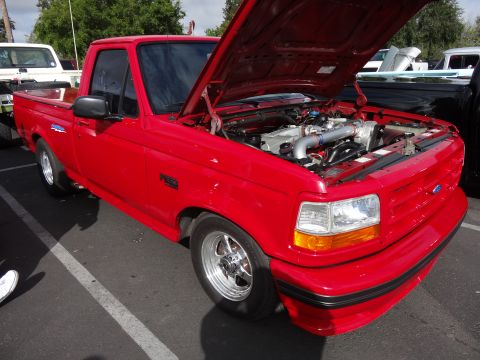 1994 Ford Bronco - This red pickup truck has its hood open, revealing the engine. The truck has chrome wheels and is parked next to another vehicle.