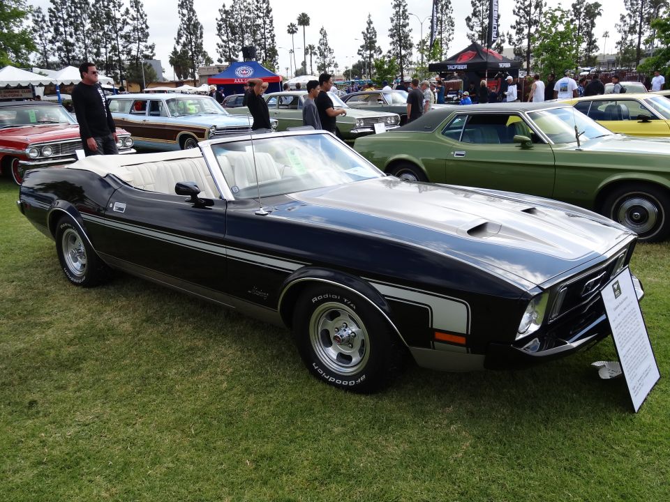 Black 1971 Ford Mustang convertible with white interior and silver racing stripes on hood and sides, parked on grass with other cars and tents in background