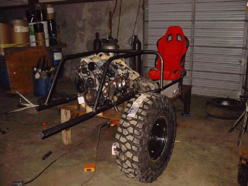 Tube-frame buggy project with a V8 engine, red racing seat, and one large mud tire in a garage