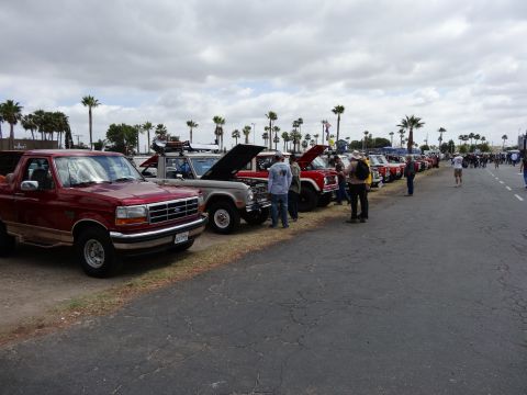 1994 Ford Bronco - A row of Ford Broncos and trucks are parked side-by-side under a cloudy sky, with several people milling around.
