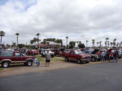 1994 Ford Bronco - A gathering of vehicles, including a maroon 1994 Ford Bronco, under a cloudy sky with palm trees.