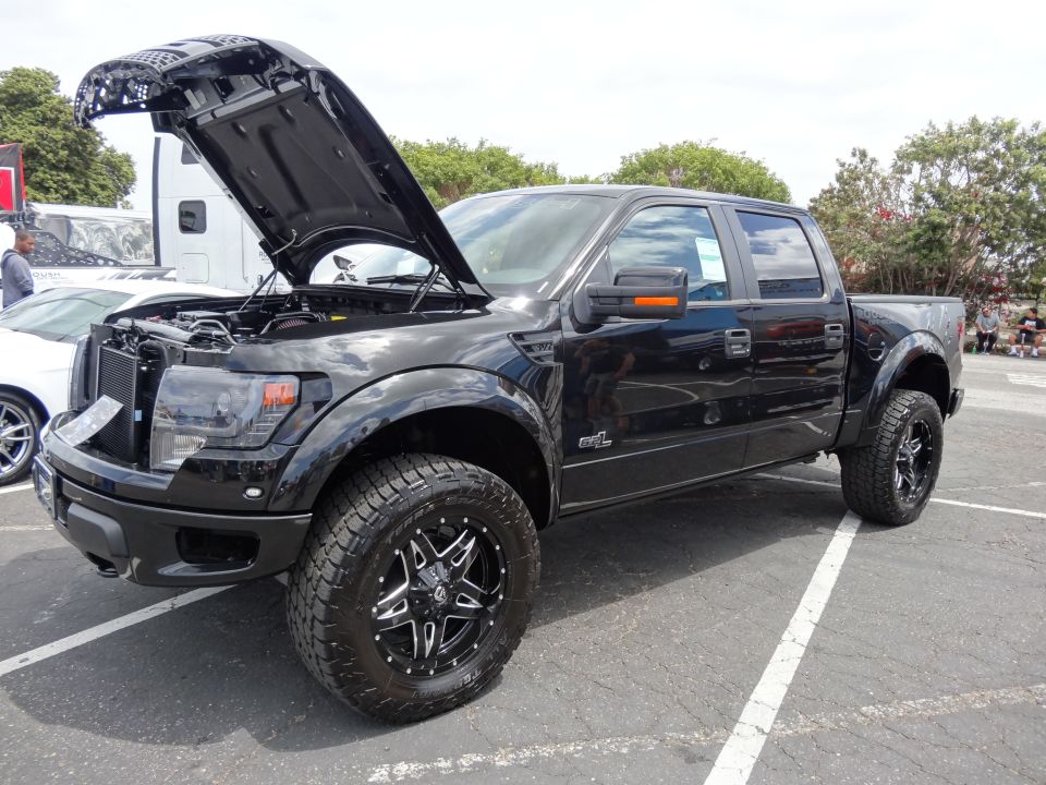 Black Ford F-150 Raptor pickup truck, hood open, showing engine bay. Aftermarket wheels and tires